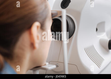 Esame dell'occhio. Bella piacevole giovane donna seduta e cercando in speciale lente pur avendo l'esame del suo occhio Foto Stock