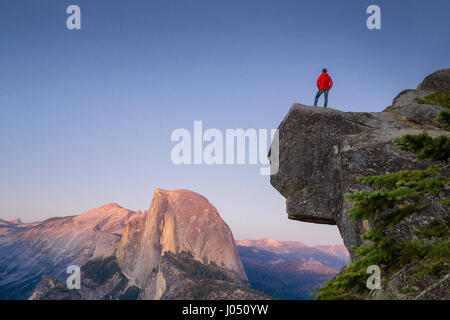Un intrepido escursionista è in piedi su una roccia a strapiombo godendo della vista verso il famoso Half Dome presso il Glacier Point si affacciano al tramonto, Yosemite NP, STATI UNITI D'AMERICA Foto Stock