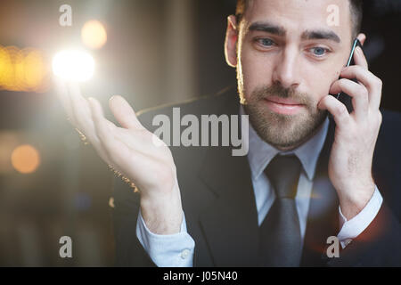 Ritratto di successo imprenditore barbuto sorridente e gesticolando mentre parla al telefono cellulare con client su sfondo nero con lens flare Foto Stock