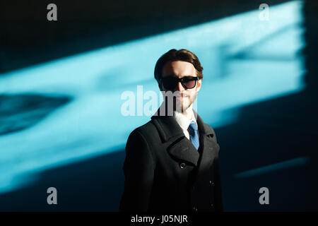 Ritratto di bello business man indossando occhiali da sole e mantello nero in condizioni di luce solare intensa contro sfondo blu e sorridente splendidamente guardando camer Foto Stock