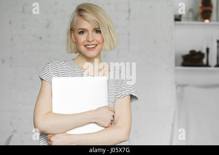 Carino giovane ragazza sorridente e detiene il portatile a luce diurna nella elegante sala. Foto Stock