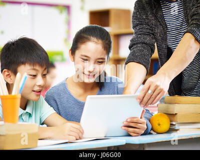 Due asian bambini di scuola elementare con tavoletta digitale con guida dal docente tutor in aula. Foto Stock