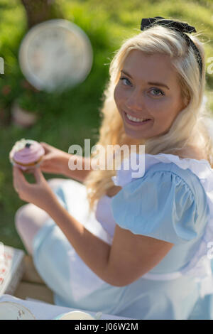 Giovane donna vestita come Alice nel Paese delle Meraviglie mantiene la torta, sorridente alla fotocamera Foto Stock