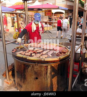 KAOHSIUNG, Taiwan -- 15 ottobre 2016: un venditore ambulante cuochi di lastre di bacon, salsicce di maiale, uova e riso in bambù su una griglia al di sopra di una grande barbec Foto Stock