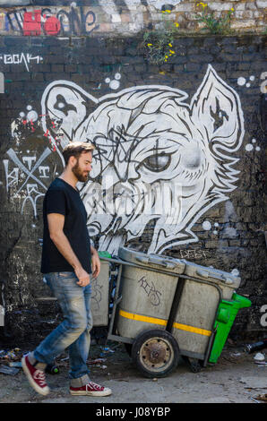 Un uomo cammina passato una strada pulitori camion e in bianco e nero dipinto murale sulla parete di un'area parcheggio off Brick Lane nella zona est di Londra. Foto Stock