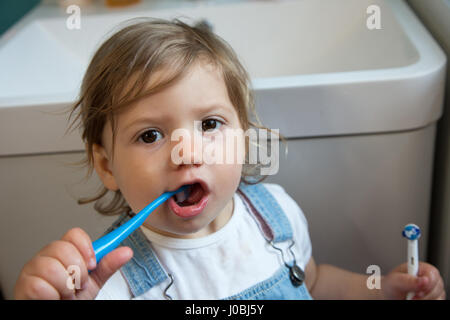 Ragazza toddler spazzolare i suoi denti utilizzando uno spazzolino da denti per bambini e tenendo un adulto spazzolino da denti elettrico nella sua altra mano. Ella è di 16 mesi Foto Stock