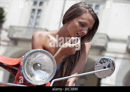 Giovane donna seduta su scooter, cercando in ala specchio, rossetto Foto Stock