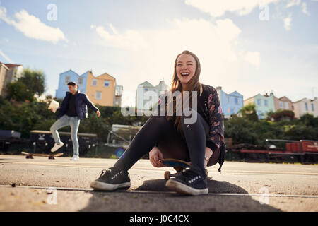 Due amici ingannare intorno all'aperto, giovane donna seduta su skateboard, ridere, Bristol, Regno Unito Foto Stock