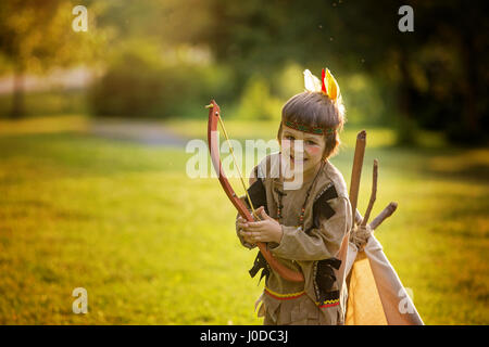 Simpatico ritratto di native american boy con costumi, giocare all'aperto nel parco con arco, frecce e ascia di guerra sul tramonto, estate Foto Stock