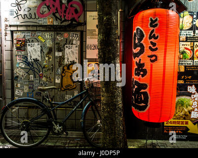 Graffiti e una bicicletta appoggiata contro un albero accanto a una grande lanterna chochin pubblicità ad un ristorante. Strada notte, Shinjuku, Tokyo, Giappone Foto Stock