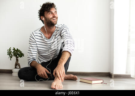 Giovane uomo spogliato camicetta ascoltando musica al suo cellulare e seduto sul pavimento accanto a un libro e un impianto Foto Stock