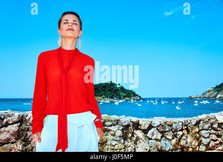 Fine settimana lussuoso rifugio. Ritratto di rilassato turistico alla moda donna in camicia rossa in piedi di fronte allo splendido scenario affacciato sulla laguna con yach Foto Stock