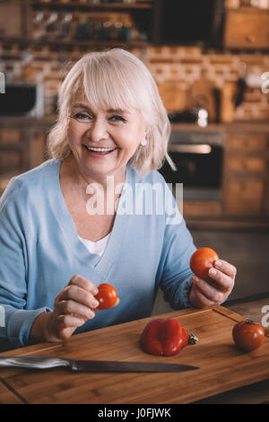 Felice donna senior tenendo i pomodori durante la cottura in cucina Foto Stock