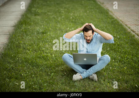 Bel giovane uomo sdraiato in erba con notebook diventa solo cattive notizie Foto Stock