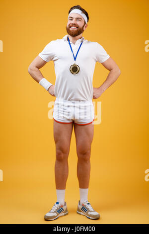 Immagine di sorridente giovane sportivo con medaglia vestite di bianco t-shirt in piedi isolato su sfondo giallo. Guardando a parte. Foto Stock