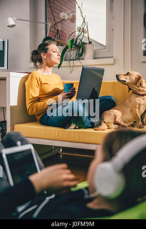Donna che indossa un maglione giallo seduto sul divano giallo utilizzando laptop e parlando con il suo cane Foto Stock