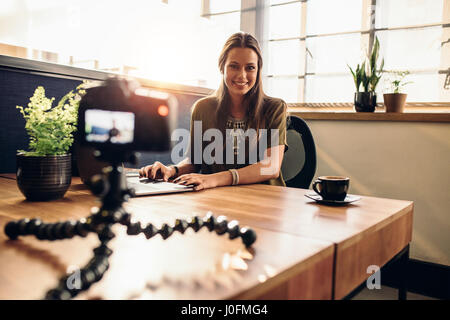 Giovane donna di registrazione di video per il suo vlog su una telecamera digitale montata su treppiede flessibile. Donna sorridente seduto alla sua scrivania che lavora su un notebook compute Foto Stock