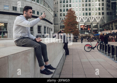 Tecnologia: un bel giovane maschio caucasico prende un selfie seduti fuori a Canary Wharf, Londra. Foto Stock