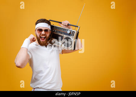 Immagine di urlando giovane uomo che indossa gli occhiali da sole azienda registratore a nastro vestite di bianco t-shirt isolate su sfondo giallo. Guardando la marca della fotocamera Foto Stock