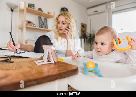 Bambino eccitato guardando smart phone mentre madre parlando su smart phone e scrivere le note a home office Foto Stock