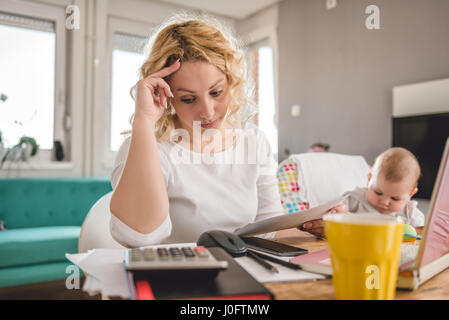 Preoccupato madre guardando la busta presso l'ufficio domestico e di prendersi cura del suo bambino Foto Stock