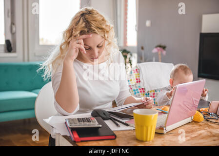 Preoccupato madre guardando la busta presso l'ufficio domestico e di prendersi cura del suo bambino Foto Stock