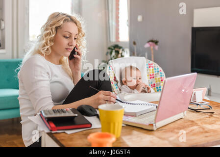 Madre di parlare su smart phone e scrivere le note a home office e di prendersi cura del suo bambino Foto Stock