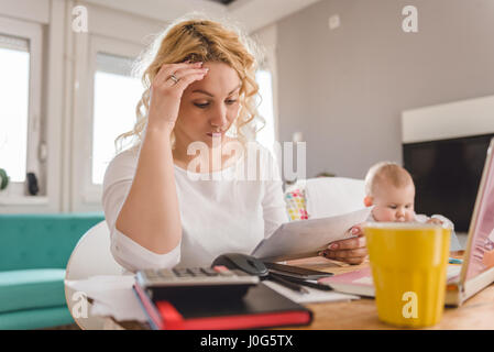 Preoccupato madre guardando la busta presso l'ufficio domestico e di prendersi cura del suo bambino Foto Stock