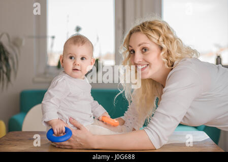 La madre e il suo bambino seduto sul tavolo con il giocattolo blu in mano e guardando la fotocamera Foto Stock