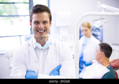 Ritratto di dentista in piedi con le braccia incrociate mentre il suo collega l'esame paziente in background Foto Stock