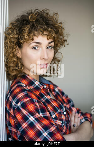 Giovani ricci naturali donna sorridente alla telecamera casual vestito con una camicia a scacchi all'interno di una casa Foto Stock