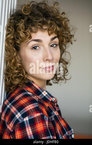 Giovani ricci naturali donna sorridente alla telecamera casual vestito con una camicia a scacchi all'interno di una casa Foto Stock