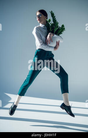 Giovane donna elegante in camicia bianca e occhiali jumping con piante verdi in vaso di fiori in mani Foto Stock