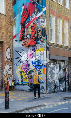 Una donna cammina davanti a una grande parete su Hanbury Street nella zona est di Londra che è coperto di arte di strada. Foto Stock