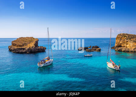 A Comino e Malta - barche a vela presso la splendida laguna blu a Comino Isola con il turchese chiaro l'acqua di mare, cielo blu e rocce in acqua Foto Stock