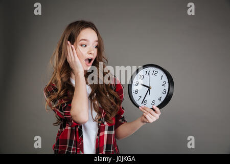 Immagine di scioccato giovane donna in piedi isolato su sfondo grigio tenendo premuto clock. Guardando a parte. Foto Stock