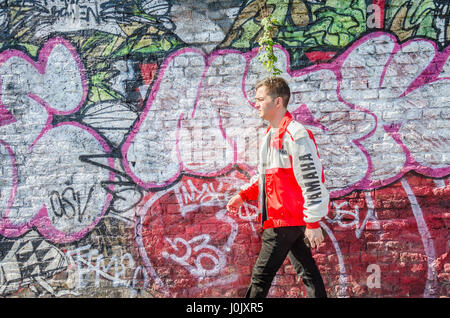 Un uomo cammina davanti ad una parete ricoperta di street art e graffiti su Brick Lane nella zona est di Londra. Foto Stock