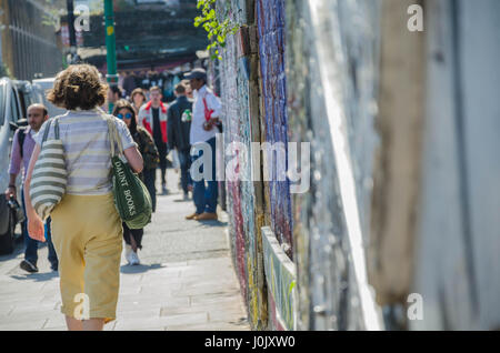 I pedoni a piedi lungo un sentiero a fianco di un muro di mattoni a Brick Lane che è coperto in strada sono e graffiti. Foto Stock