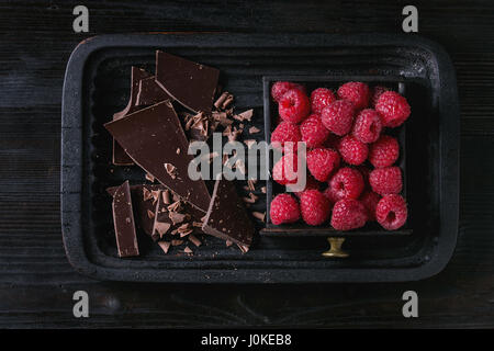 Dark e tritare il latte al cioccolato e i chip sulla vecchia piastra con lamponi freschi in black box in legno su nero bruciato sullo sfondo di legno. Vista da sopra con spa Foto Stock