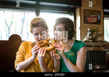 Due amiche mangiando pretzel, guardando la telecamera impostazione indoor. Entrambi sono sorridente e godendo il loro tempo insieme Foto Stock