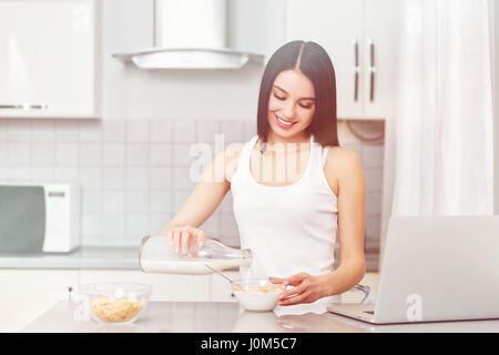 Una giovane donna si prepara un basso contenuto calorico prima colazione su una prescrizione mentre guardando il suo computer portatile. La dieta, mele, muesli, fiocchi di mais - il concetto di hea Foto Stock