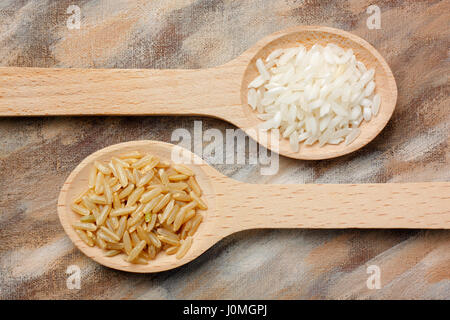 Due cucchiai di legno con bianco e marrone di grani di riso su sfondo dipinto Foto Stock