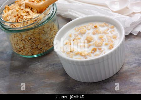 Ramekin riempito da porridge con il latte e il vasetto di vetro con le materie di fiocchi d'avena e cucchiaio di legno Foto Stock