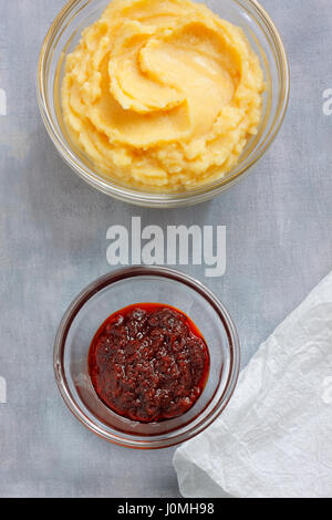 Hummus e salsa Harissa in ciotole in vetro su verniciato grigio Sfondo tessile con un panno di carta. Vista aerea. Foto Stock