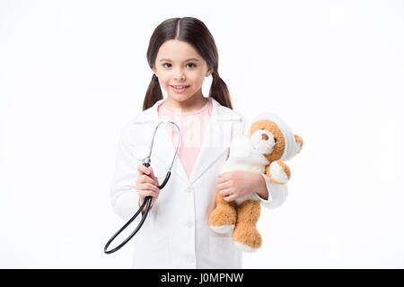 Sorridente bambina in medico uniforme tenendo bendato orsacchiotto e stetoscopio Foto Stock