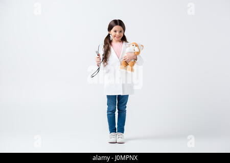 Sorridente bambina in medico uniforme tenendo bendato orsacchiotto e stetoscopio Foto Stock