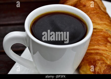 Close-up di una tazza di caffè nero e un croissant su una tavola di legno, all'esterno. Foto Stock