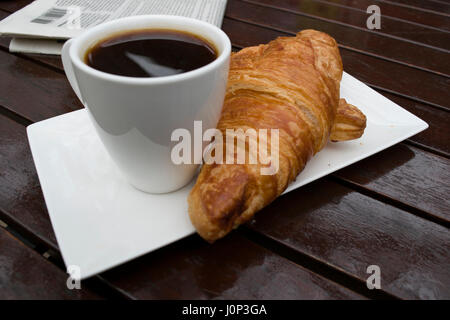 Una tazza di caffè nero e un croissant francesi, un giornale su un tavolo di legno, all'esterno. Foto Stock