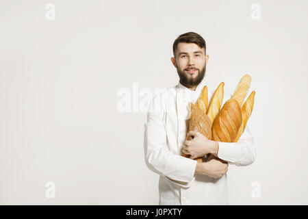 Bello baker in uniforme tenendo baguette di pane con ripiani su uno sfondo bianco. Uomo bello tenendo il pane caldo nelle sue mani su bianco backgrou Foto Stock