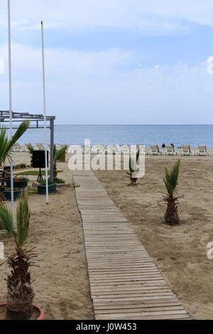 Percorso di legno al mare con piccole palme si trova lungo e lettini sulla spiaggia di sfondo Foto Stock